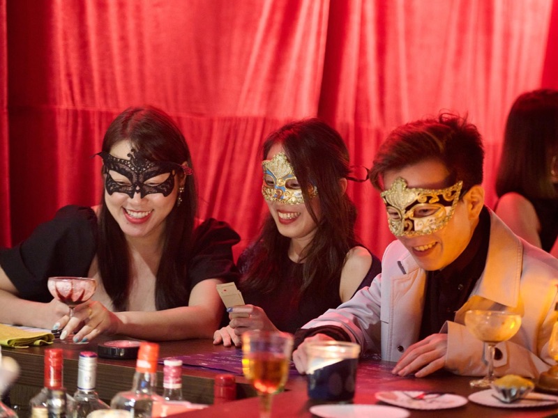 Tiga tamu bertopeng tertawa di meja bar bertirai merah